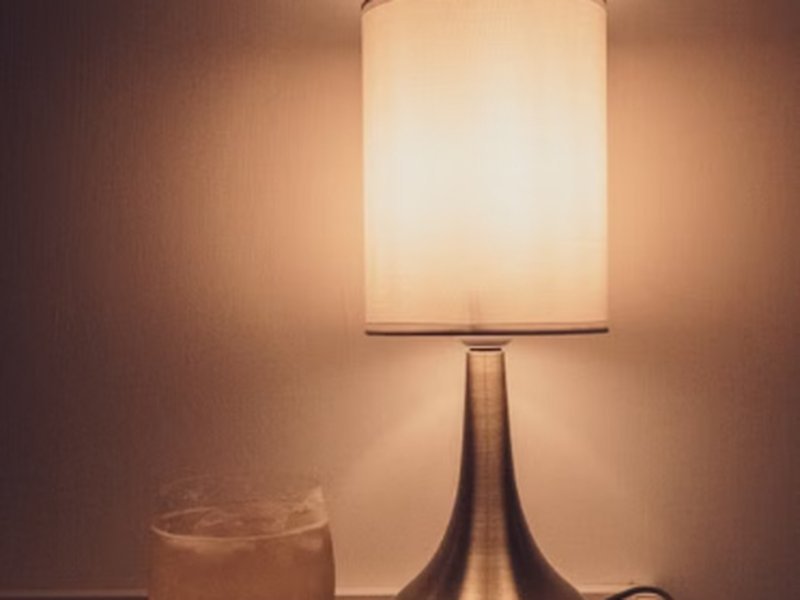 Luminária de mesa acesa com luz quente sobre criado-mudo ao lado da cama à noite